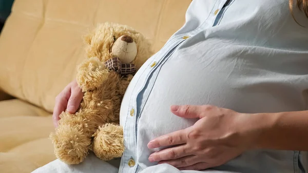 인지할 수없는 임신부가 소파에 앉아 곰 장난감을 들고 배를 저으는 모습 — 스톡 사진