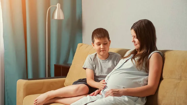 Pregnant mom and son on the couch. Boy stroking and hugging belly — Stock Photo, Image