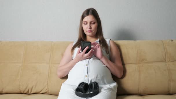 Mujer embarazada con teléfono poniéndose auriculares mientras está sentada en el sofá — Vídeos de Stock