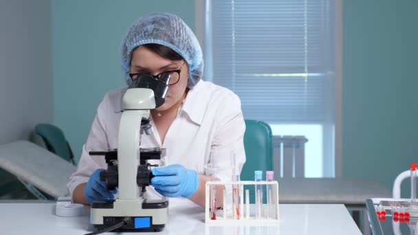 Mulher jovem olha para a amostra através do microscópio na clínica — Vídeo de Stock