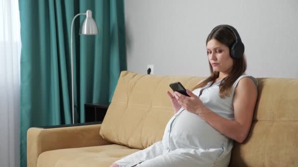 Беременная женщина в наушниках смотрит в телефон, сидя на диване — стоковое видео