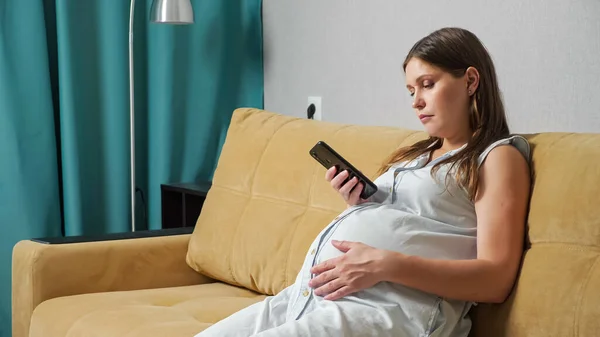 Young pregnant woman typing text on the phone and stroking belly while sitting on the couch — Stock Photo, Image