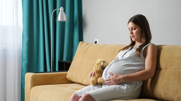 Jeune femme enceinte caressant ventre tenant ours jouet tout en étant assis sur le canapé — Photo