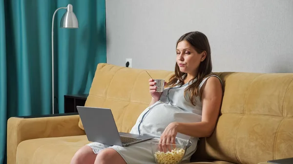Pregnant woman eating yogurt and popcorn and looking at laptop while sitting on sofa — Stock Photo, Image