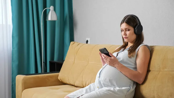 Pregnant woman with headphones looking at the phone while sitting on the couch — Stock Photo, Image