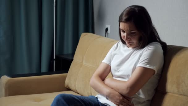 Fiatal nő görnyedt át tartja hasát, miközben ül a kanapén — Stock videók