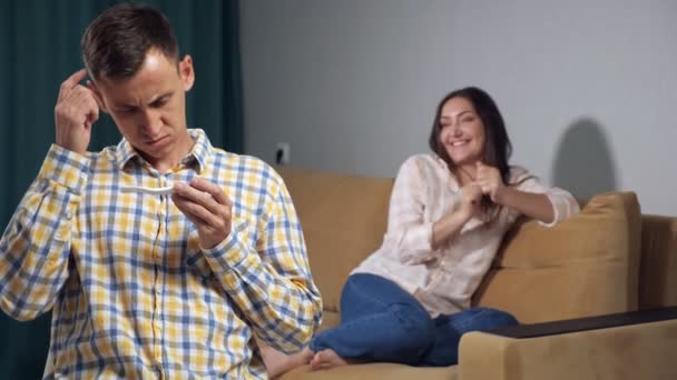 Fiatal férfi zavarodottság néz egy terhességi teszt hátterében egy boldog terhes nő, lassított felvétel — Stock videók