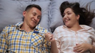 Erkek ve kadın yatakta yatarken pozitif gebelik testine sevinirler.