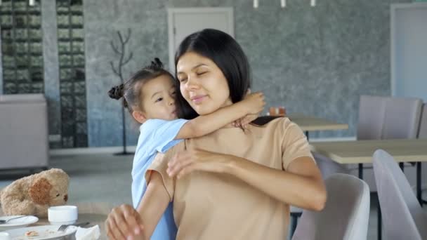 Bambina abbraccia mamma bruna con sorriso vicino al tavolo — Video Stock