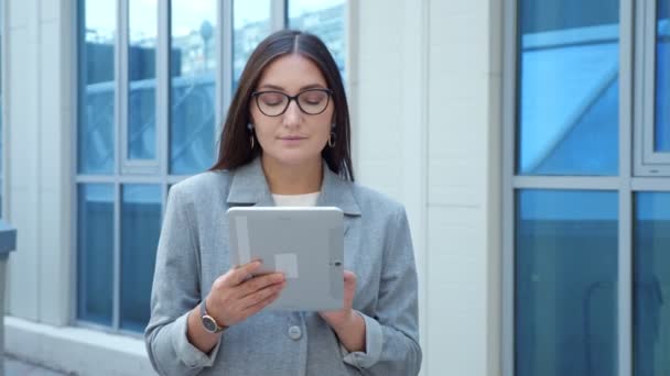 Woman in a formal suit and glasses looks into a tablet on the street, slow motion — ストック動画