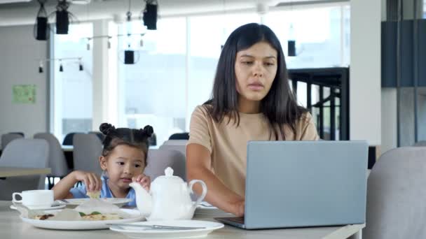 Aziatisch zakenvrouw mam werkt zitten aan tafel met thee en grijs laptop en kind eet pizza slice naar peuter — Stockvideo