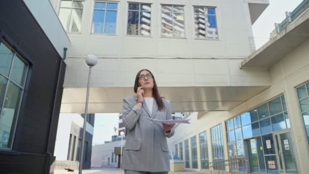 Kvinde i et formelt jakkesæt taler i telefon og ser på papirer gå ned ad gaden – Stock-video