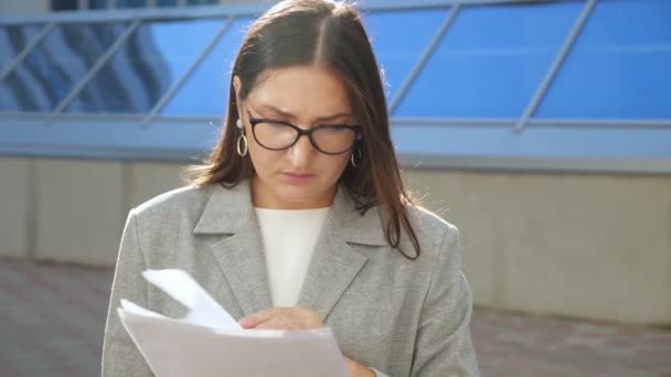 Fiatal nő, öltönyben és szemüvegben, komoly arccal vizsgálja a dokumentumokat az utcán — Stock videók