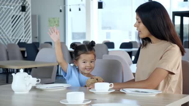 Småbarn flicka höjer handen med leende sitter vid mamma i caféet — Stockvideo