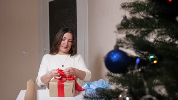 Ung kvinna dekorerar låda med rött band bredvid julgran — Stockvideo