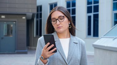 Gözlüklü ve takım elbiseli genç bir kadın binaların arasında telefona bakarak yürüyor.