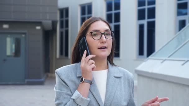 Mujer joven en gafas y un traje está caminando entre edificios y hablando por teléfono, cámara lenta — Vídeos de Stock