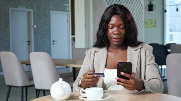 Mujer negra paga en línea con el teléfono sentado en la mesa en la cafetería — Vídeos de Stock