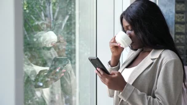 Mujer afroamericana bebe café y mensajes de texto por teléfono — Vídeos de Stock