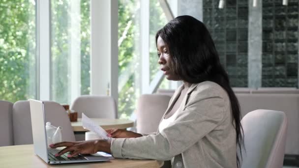 Afro-americká dáma typy na notebooku čtení papíry v kavárně — Stock video