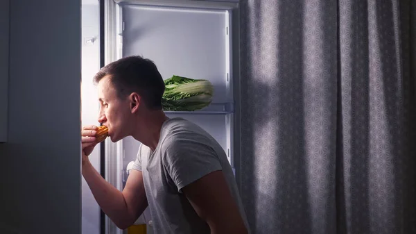 Человек открывает дверь холодильника и тайно ест эклер на тёмной кухне. — стоковое фото