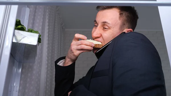 Мужчина разговаривает по телефону, ест сэндвич с маслом возле холодильника — стоковое фото