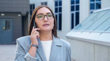 Gözlüklü ve takım elbiseli genç bir kadın binaların arasında yürüyor ve telefonla konuşuyor.