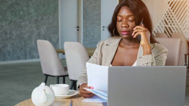 Kâğıtları olan Afro-Amerikalı bir kadın kafede telefonla konuşuyor.