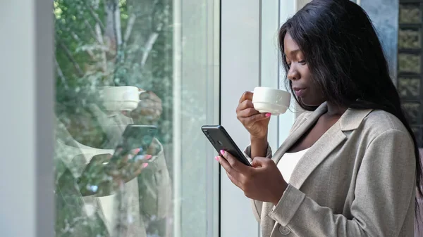 Mujer afroamericana bebe café y mensajes de texto por teléfono —  Fotos de Stock