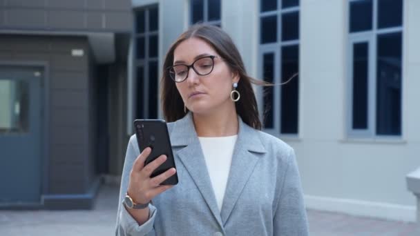 Mujer joven en gafas y un traje camina entre edificios mirando el teléfono, cámara lenta — Vídeos de Stock