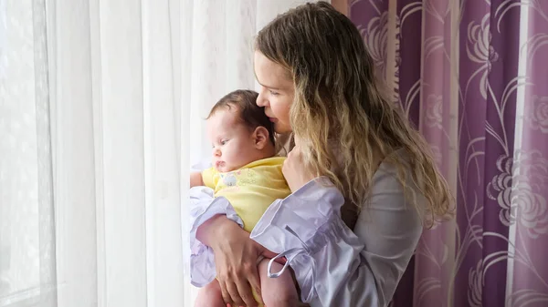 Jeune femme tenant un petit enfant dans les bras tout en se tenant par la fenêtre — Photo