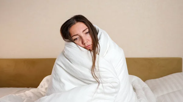 Mujer enferma envuelta con edredón blanco suave se sienta en el dormitorio — Foto de Stock
