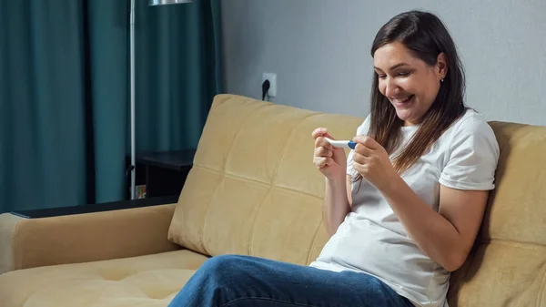 Happy woman holding pregnancy test in hands sitting on sofa — Stock Photo, Image