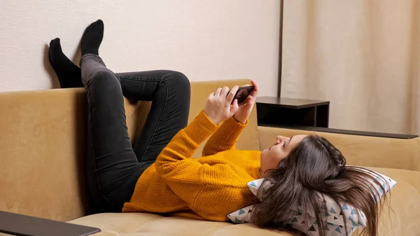 Long haired brunette in pullover on sofa types on smartphone — Stock Photo, Image