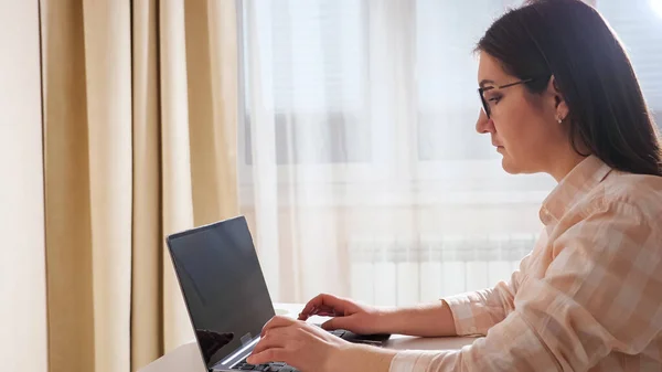 Жінка сидить за столом і друкує на ноутбуці на задньому плані вікна. — стокове фото