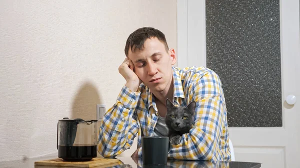 Sleepy hombre se sienta con gris gato en la mesa en la cocina — Foto de Stock