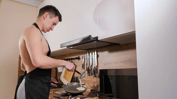 Hombre con un torso desnudo y en un delantal negro prepara panqueques en la cocina —  Fotos de Stock