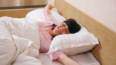 Kadın uyku maskesini çıkarır, kollarını uzatır ve yatağa uzanır.