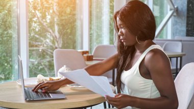 Afro-Amerikalı kadın dizüstü bilgisayarla çalışan kağıtlara bakıyor.