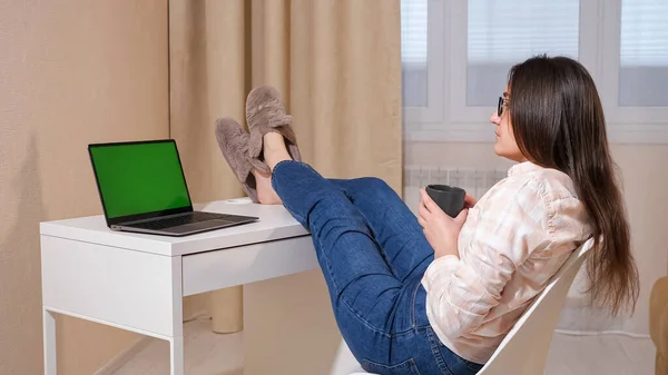 Jovem mulher joga as pernas na mesa e bebe café enquanto olha para o laptop com tela verde — Fotografia de Stock