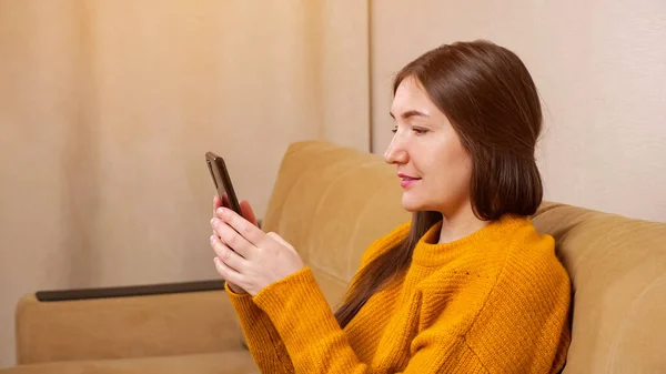 Brunette in pullover sits on sofa and types on smartphone — Stock Photo, Image