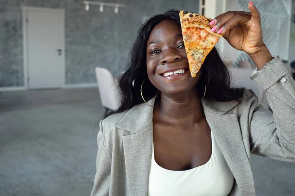 Jovem afro-americana em terno de negócios sorrindo, cobrindo o olho com uma fatia de pizza — Fotografia de Stock