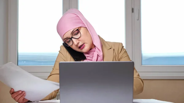 Kvinna i hijab undersöker dokument och fungerar på bärbar dator när du pratar i telefon — Stockfoto