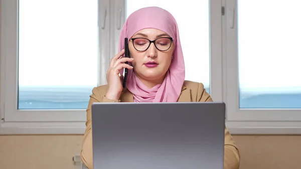 Muselman kvinna i rosa slöja och glasögon talar på telefonen medan sitter vid bordet med bärbar dator — Stockfoto