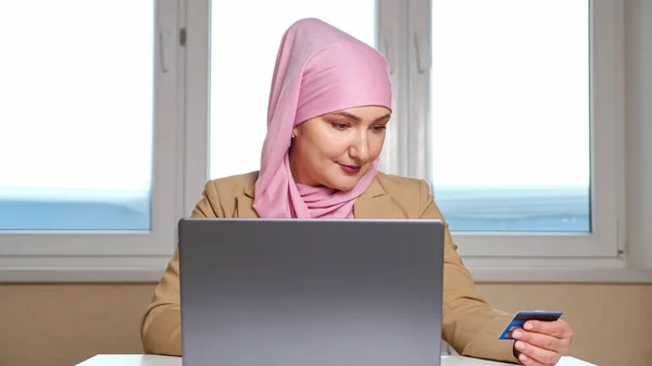 Νεαρή Μουσουλμάνα με ροζ μαντίλα κάνει ηλεκτρονικές αγορές σε φορητό υπολογιστή κρατώντας μια τραπεζική κάρτα στα χέρια — Φωτογραφία Αρχείου