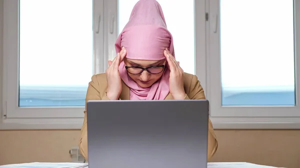 Frau im Hidschab hält ihren Kopf und massiert Schläfen, während sie an einem Laptop arbeitet — Stockfoto