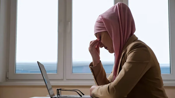 Kvinna i rosa hijab tar en paus och masserar näsryggen medan du arbetar på en bärbar dator — Stockfoto