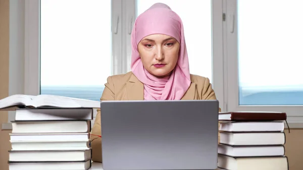 Γυναίκα με μαντίλα δακτυλογραφεί σε φορητό υπολογιστή ανάμεσα σε στοίβες βιβλίων — Φωτογραφία Αρχείου