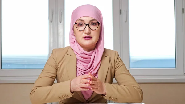 Femme en hijab et lunettes s'assoit à la table et parle à la caméra, zoomant hors de la caméra — Photo