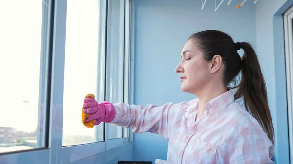 Mladá žena v rukavicích se sprejem a hadrem myje okno — Stock fotografie
